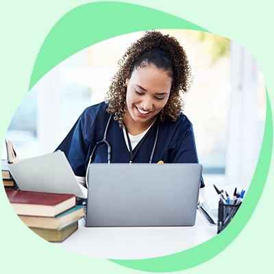 Enfermera alegre usando una laptop, sentada en un escritorio con libros y utensilios de escritura